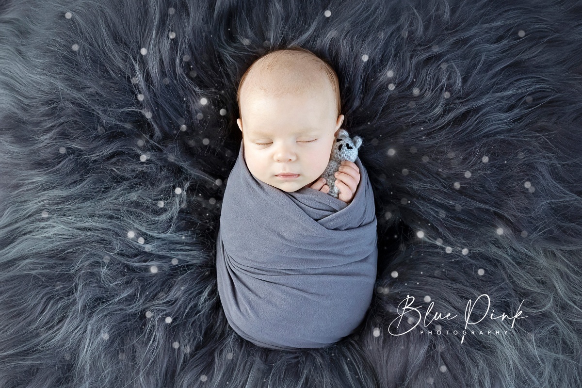 A newborn baby is wrapped in a snug potato shape using a grey elastic wrap, holding a small teddy bear in his hands while laying on a soft grey natural fur. 